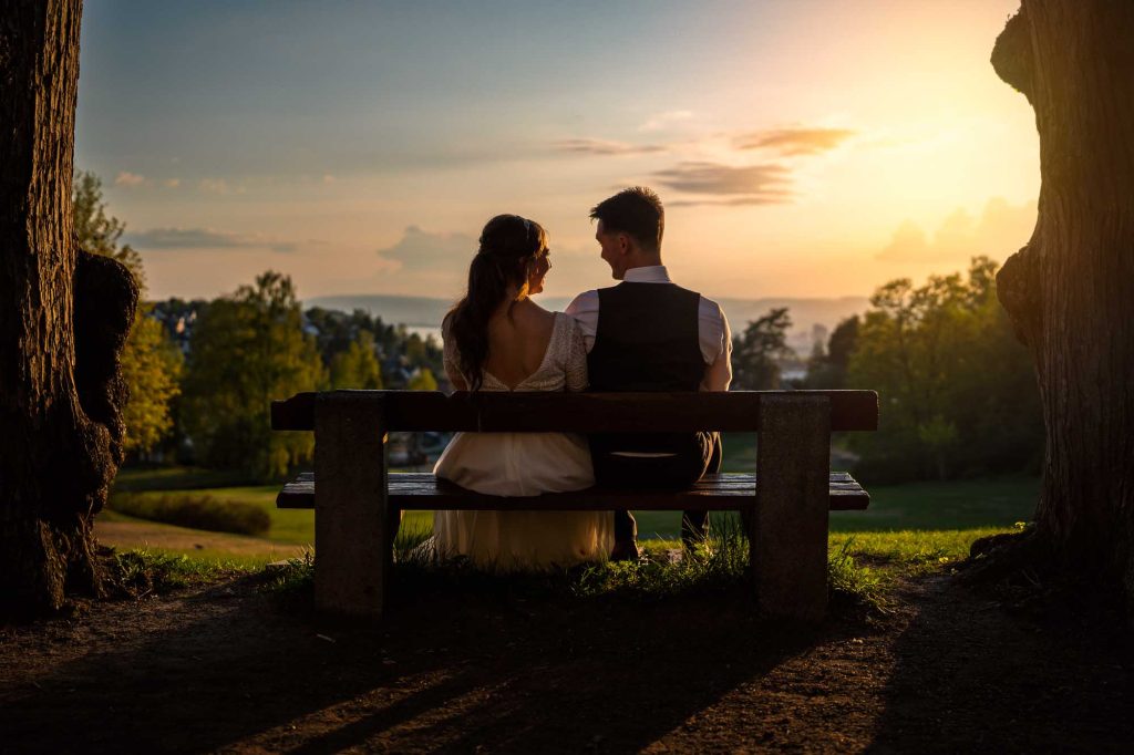 Bröllopspar sitter bredvid varandra på bänk i solnedgången.